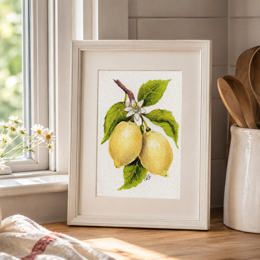 A framed watercolor print of two yellow lemons on a branch with green leaves and a small white flower, displayed on a kitchen counter next to a window, daisies, and wooden utensils.