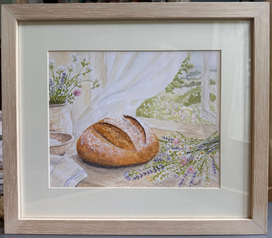 Framed artwork of a loaf of bread on a table with flowers and a window in the background.