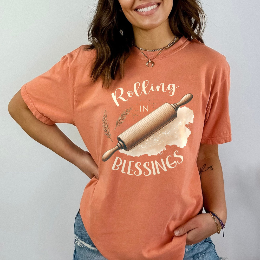 Person wearing a peach-colored t-shirt with a graphic and text 'Rolling in Blessings' on a plain background