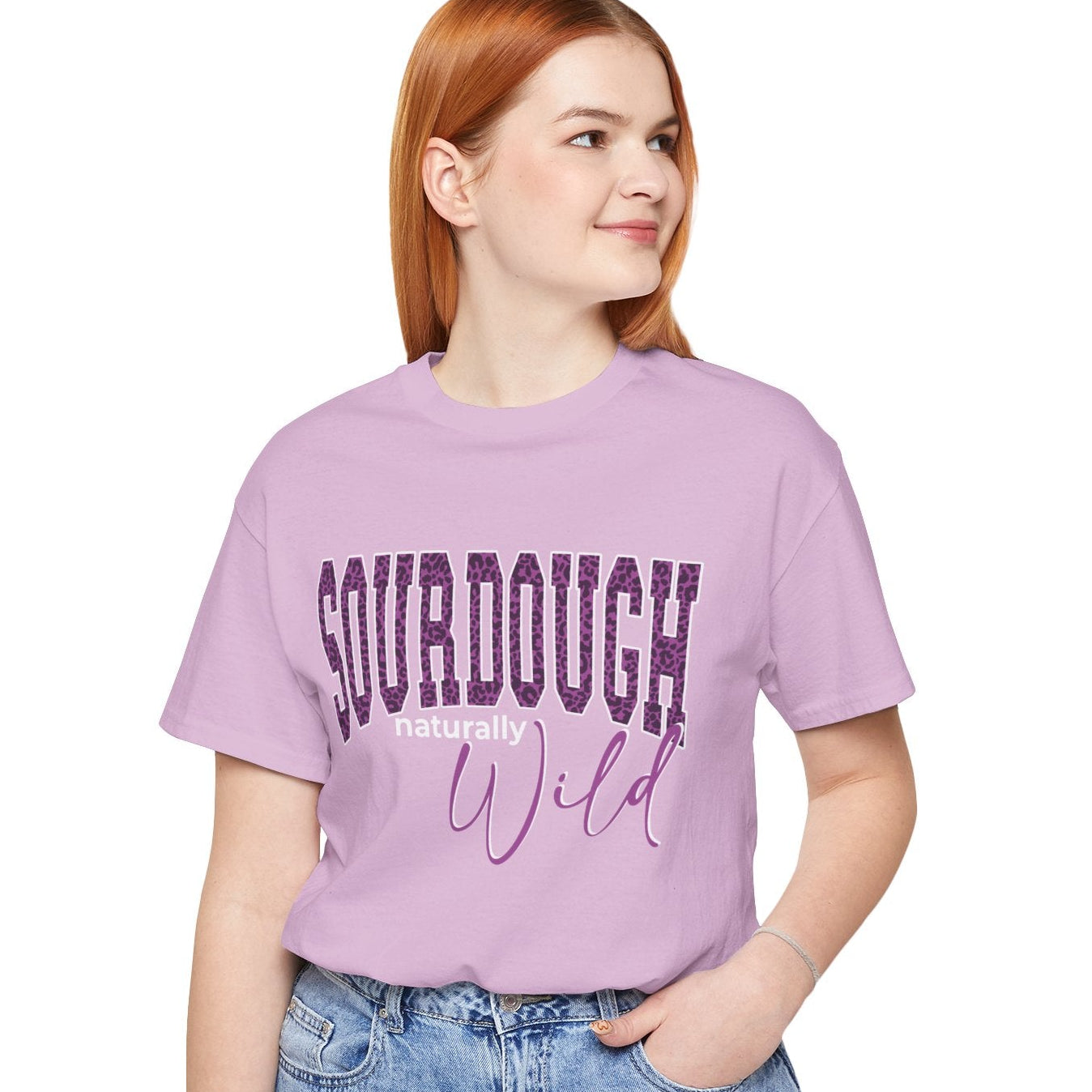 A woman with red hair wearing a lavender purple t-shirt with 'SOURDOUGH baking Wild' printed on the front, paired with light blue jeans, against a plain white background.