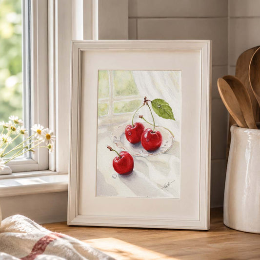 A framed still life painting of three red cherries on a white tablecloth, displayed on a wooden countertop in a bright kitchen setting next to a window and wooden utensils.