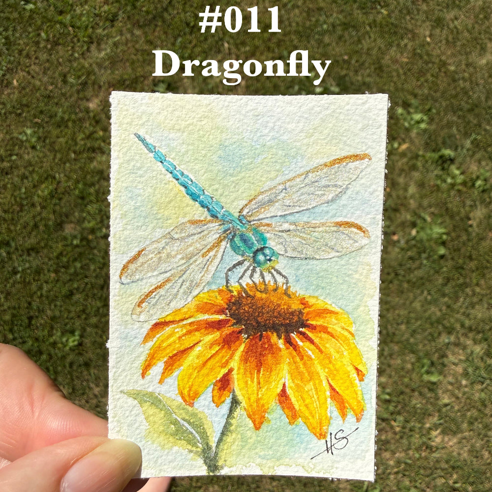 A hand holds a small 2.5 x 3.5 watercolor painting of a blue-green dragonfly perched on a yellow-orange flower, photographed outdoors on grass. White text at the top reads '#011 Dragonfly'.