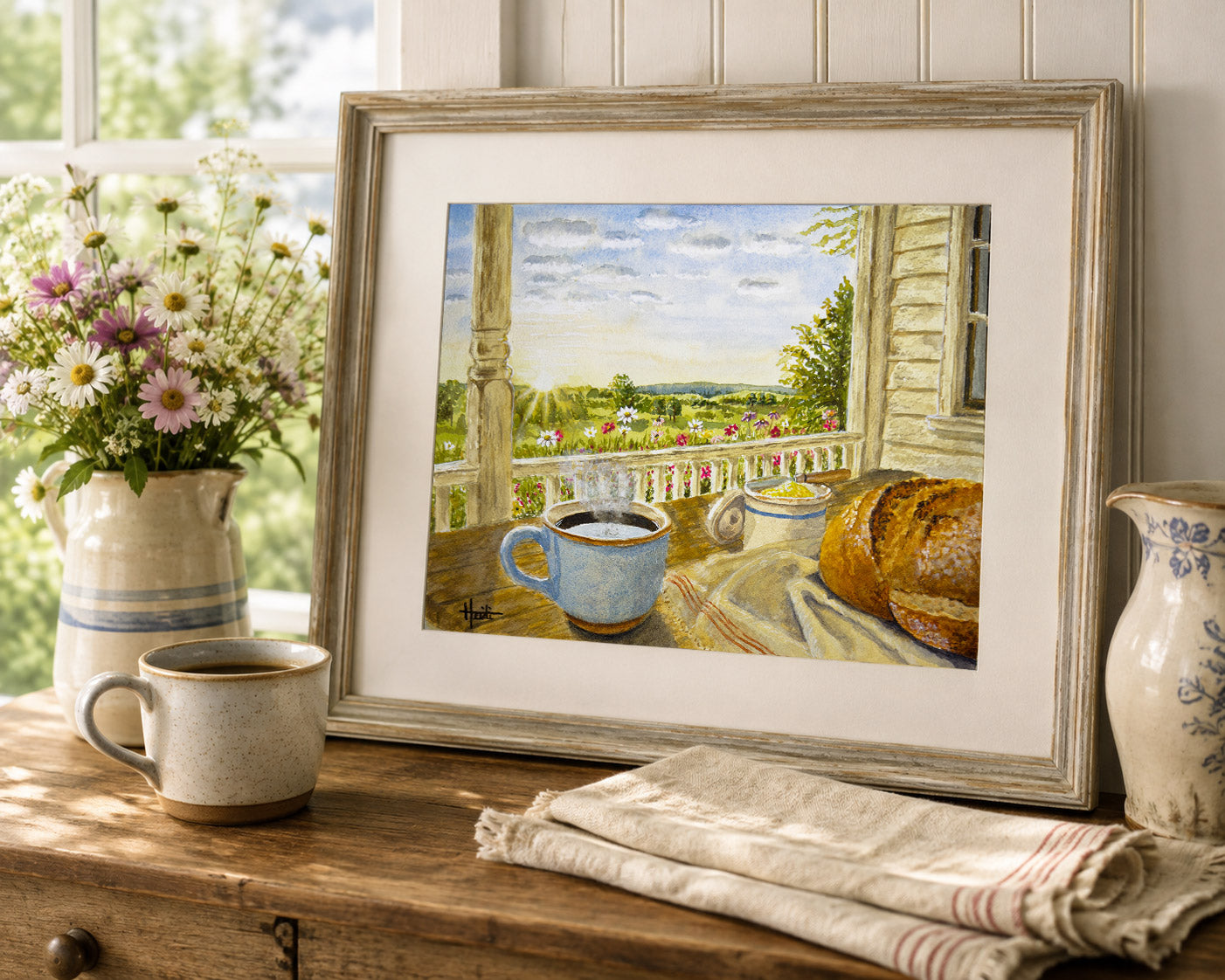 A framed art print depicting a cozy porch scene with a blue mug, bread, flowers, and a landscape under a cloudy sky, styled on a wooden table with a mug, flowers, and a cloth napkin.