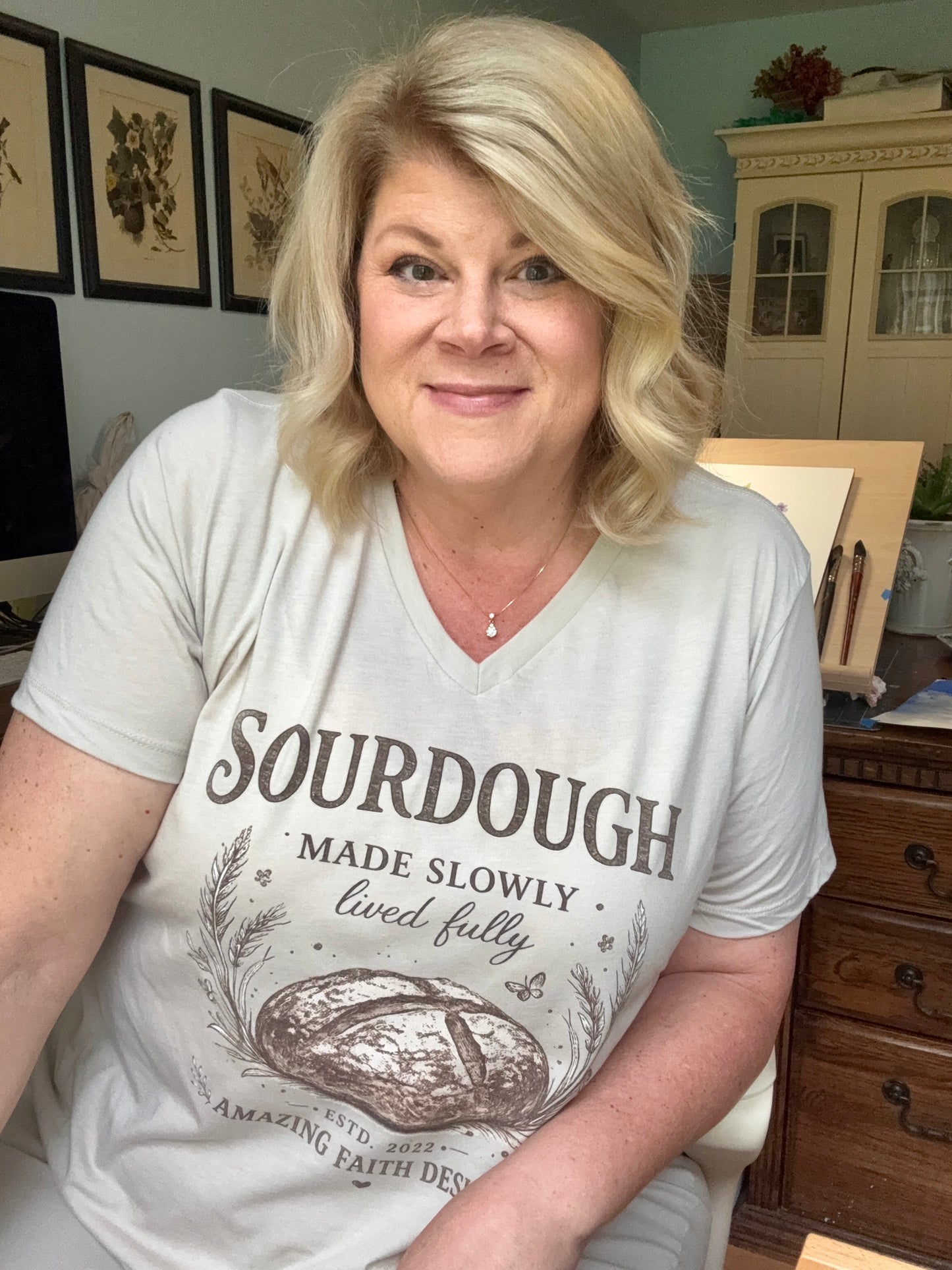 Woman wearing a heather dust t-shirt with 'Sourdough Made Slowly, Lived Fully' text and bread graphic in a home setting.
