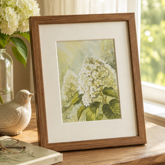 A framed botanical art print of white hydrangeas in a warm brown wood frame, styled on a wooden table next to a vase of flowers, a small bird figurine, and a book in a bright room.