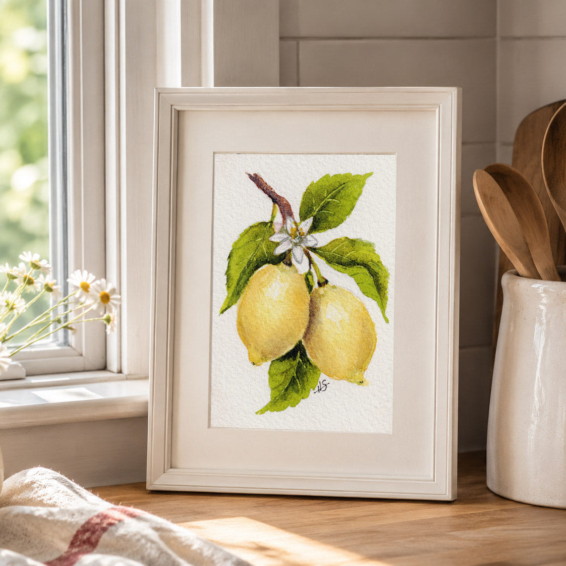 A framed watercolor print of two yellow lemons on a branch with green leaves and a small white flower, displayed on a kitchen counter next to a window, daisies, and wooden utensils.