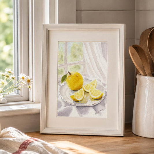 A framed watercolor painting of a yellow lemon and lemon slices on a plate by a window, styled on a kitchen counter next to a vase of daisies and wooden utensils.