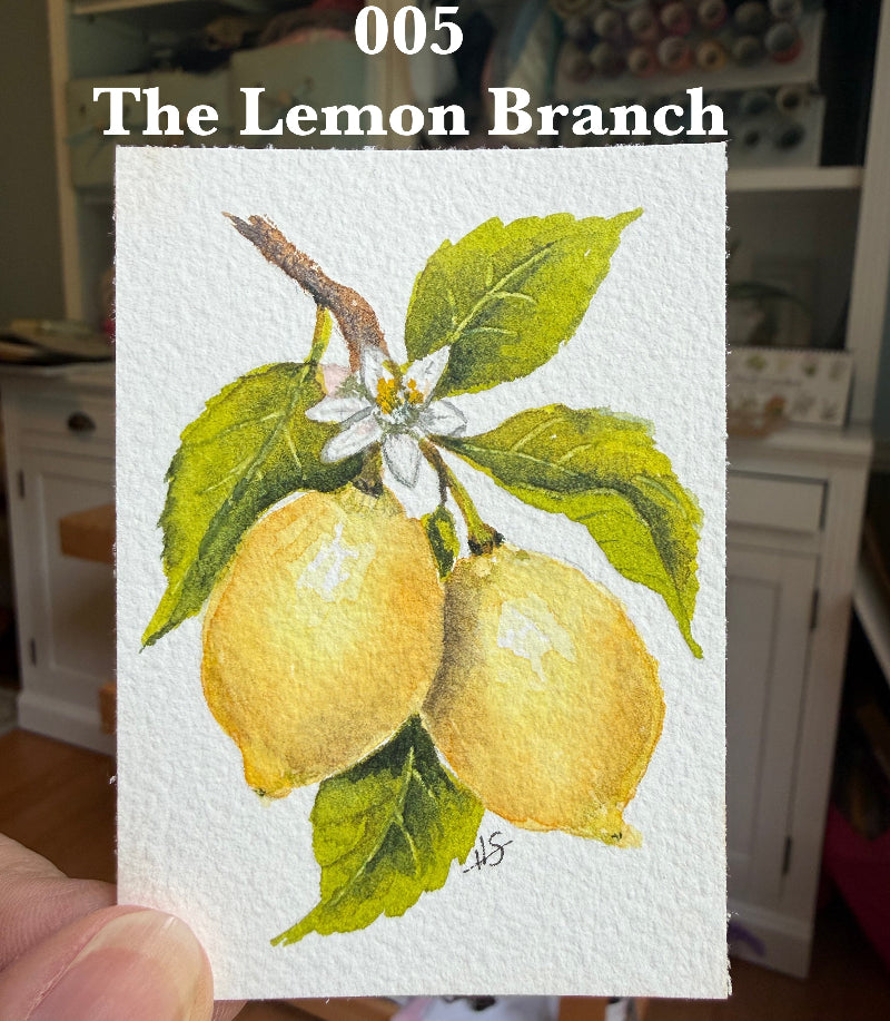 A hand holds a small watercolor painting (ACEO) of two yellow lemons on a branch with green leaves and a white flower, with the text '005 The Lemon Branch' above it. The background shows a real indoor room with shelves and furniture.
