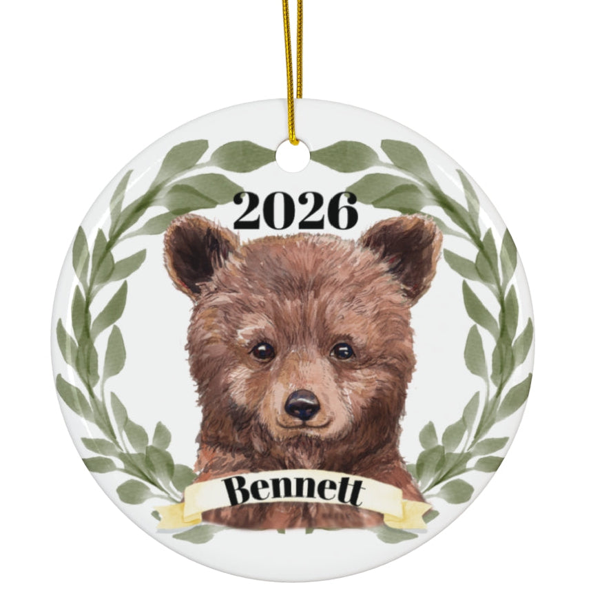 A round ceramic ornament featuring a watercolor illustration of a brown bear cub framed by green leaves, with the text '2026' above and a banner reading 'Bennett' below. The ornament hangs from a gold string against a plain white background.