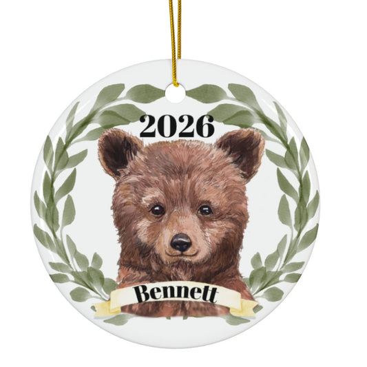 A round ceramic ornament featuring a watercolor illustration of a brown bear cub framed by green leaves, with the text '2026' above and a banner reading 'Bennett' below. The ornament hangs from a gold string against a plain white background.