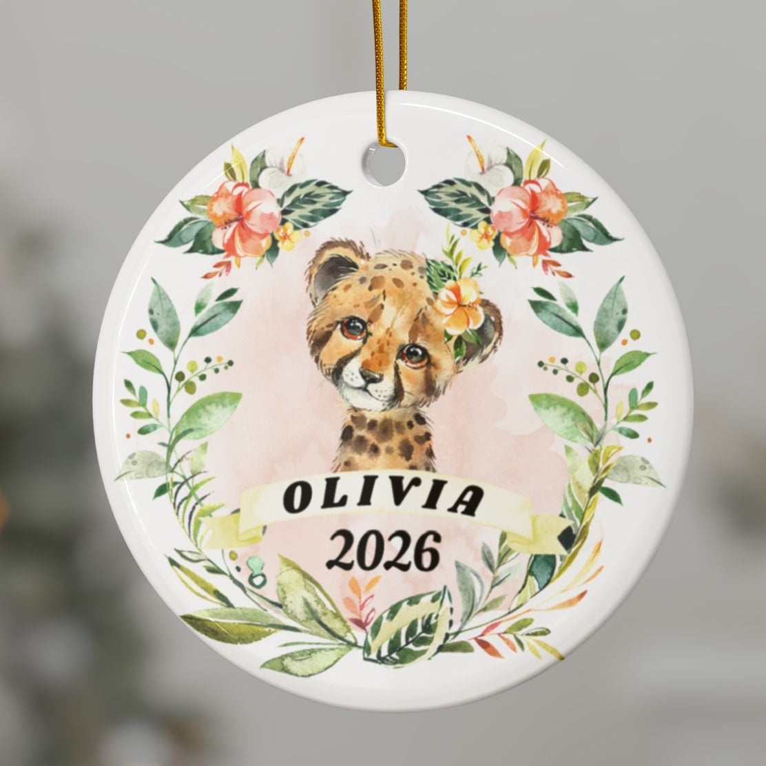 A personalized round Christmas ornament featuring a watercolor cheetah cub surrounded by floral greenery, hanging from a gold string on a Christmas tree branch. The ornament is customized with the name 'OLIVIA' and the year '2026' in black text.