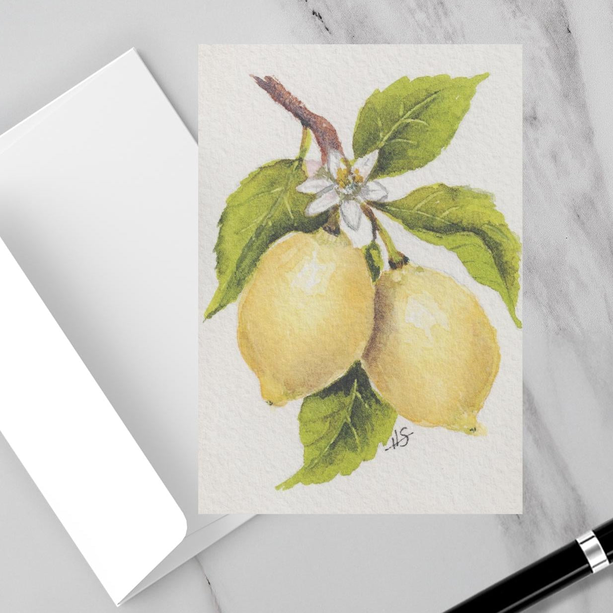 A flat-lay arrangement on a white marble surface featuring a small art print of two yellow lemons with green leaves, a black pair of eyeglasses, a white notebook, and a black pen.