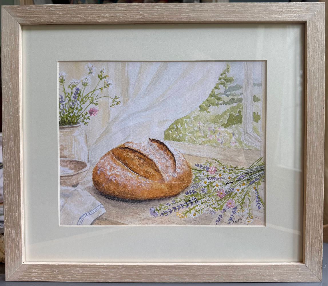 Framed artwork of a loaf of bread on a table with flowers and a window in the background.
