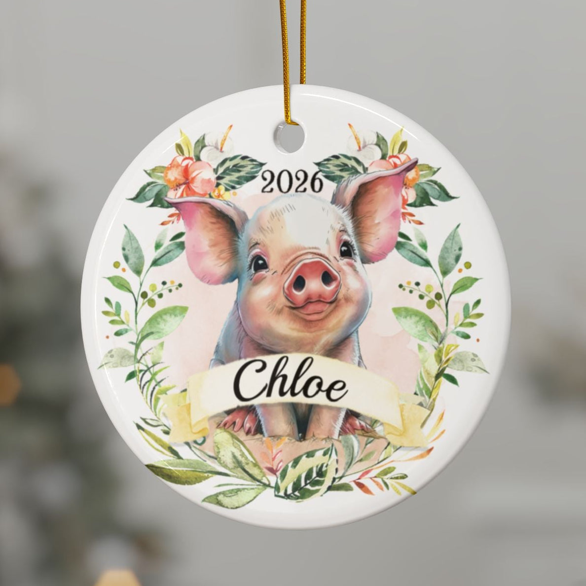 A personalized round Christmas ornament featuring a watercolor illustration of a pig with the name 'Chloe' and the year '2026', hanging from a gold string on a Christmas tree branch.