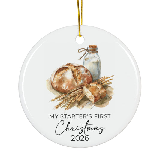 A round white ceramic Christmas ornament with a gold hanging string, featuring a watercolor illustration of bread, wheat, and a glass bottle, with the text 'MY STARTER'S FIRST Christmas 2026' printed below.